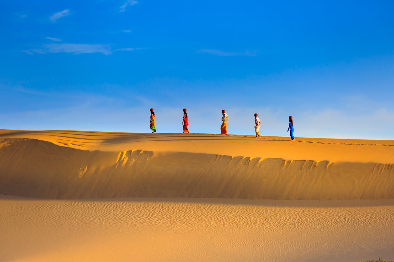 A group of people walking across golden sand dunes under a clear blue sky on a sunny day.